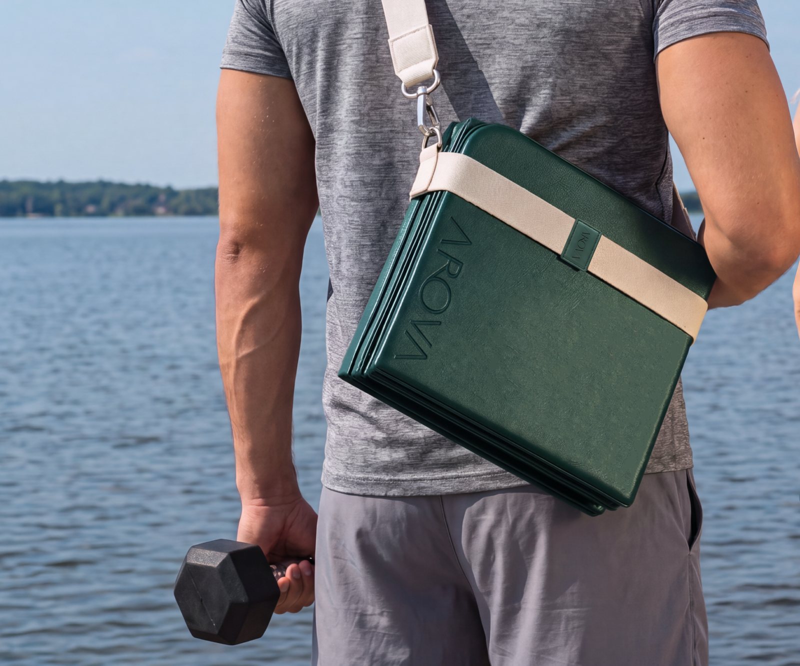 A man by the water holding a dumbbell, the folded AROVA mat across his back on its strap